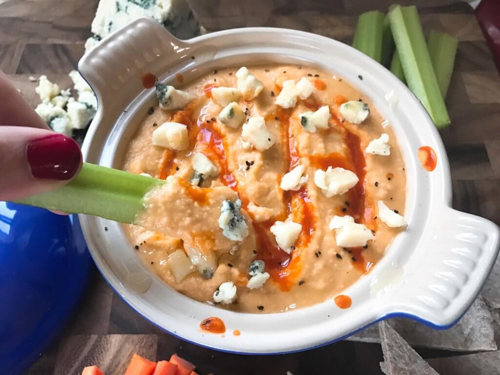 A hand dunking celery into a bowl of Buffalo Wing Hummus