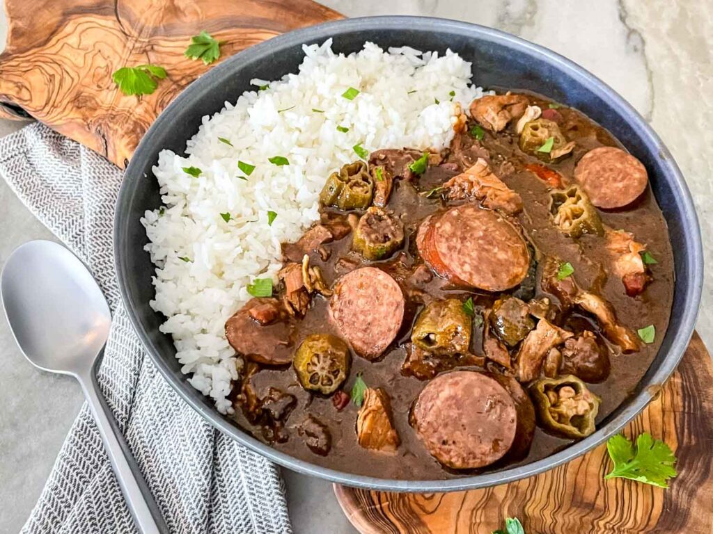 Authentic Chicken and Smoked Sausage Gumbo in a gray bowl with rice sitting on a cutting board