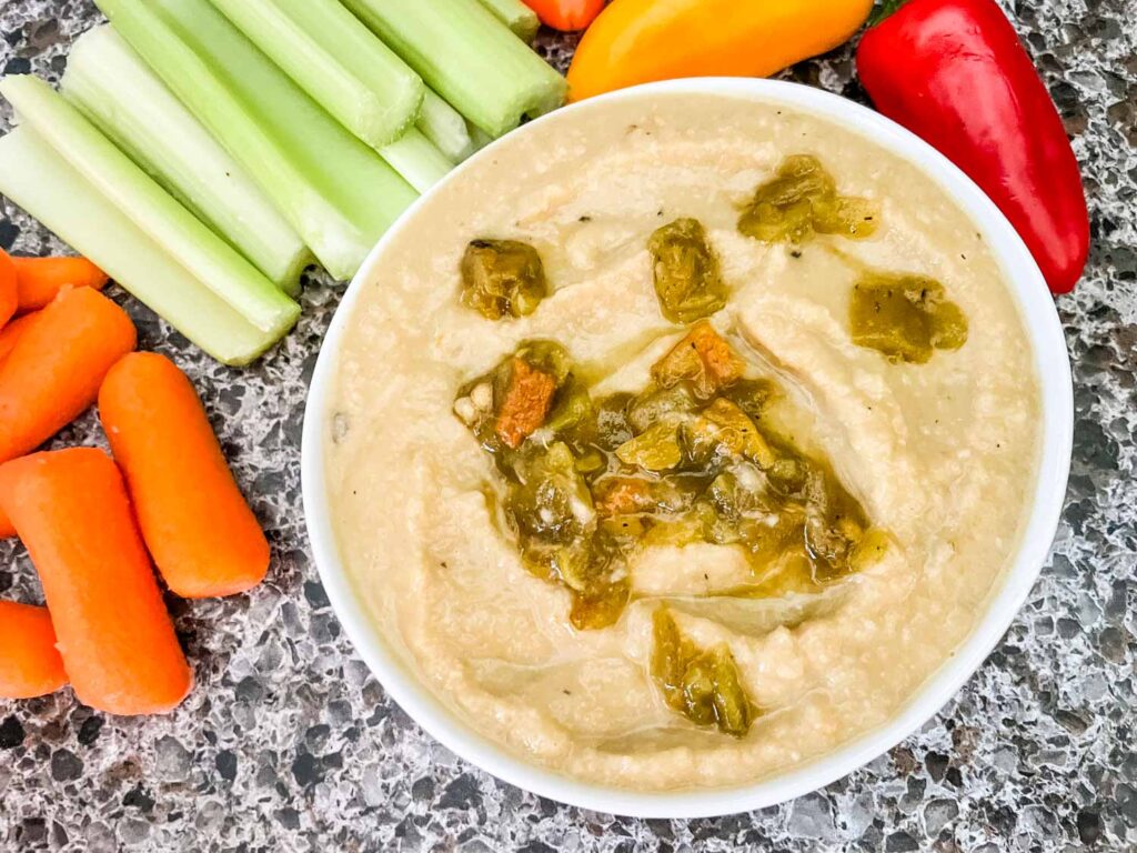 Top view of Roasted Hatch Green Chile Hummus in a small white bowl with peppers garnished on top. Sitting on a speckled gray counter with celery and carrots around it.