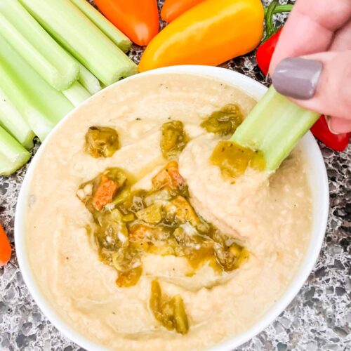 Top view of Roasted Hatch Green Chile Hummus in a small white bowl with peppers garnished on top. Sitting on a speckled gray counter with celery and carrots around it. A hand is dipping a piece of celery into the hummus.