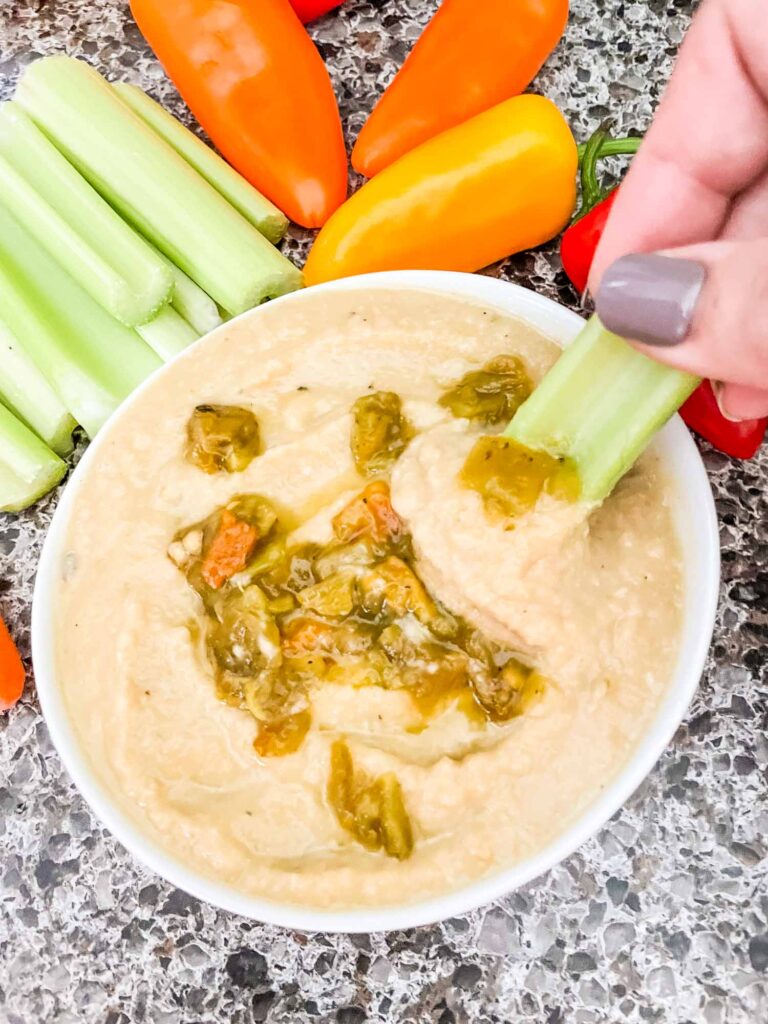 Top view of Roasted Hatch Green Chile Hummus in a small white bowl with peppers garnished on top. Sitting on a speckled gray counter with celery and carrots around it. A hand is dipping a piece of celery into the hummus.