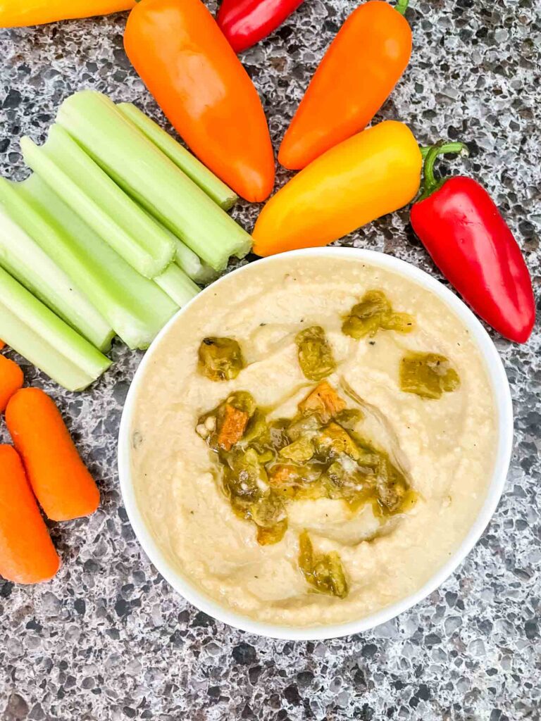 Top view of Roasted Hatch Green Chile Hummus in a small white bowl with peppers garnished on top. Sitting on a speckled gray counter with celery, peppers, and carrots around it.
