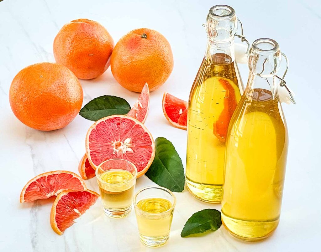 Two bottles of Grapefruit Limoncello (Grapefruit-cello) with two shots of it on a counter surrounded by fresh grapefruits.