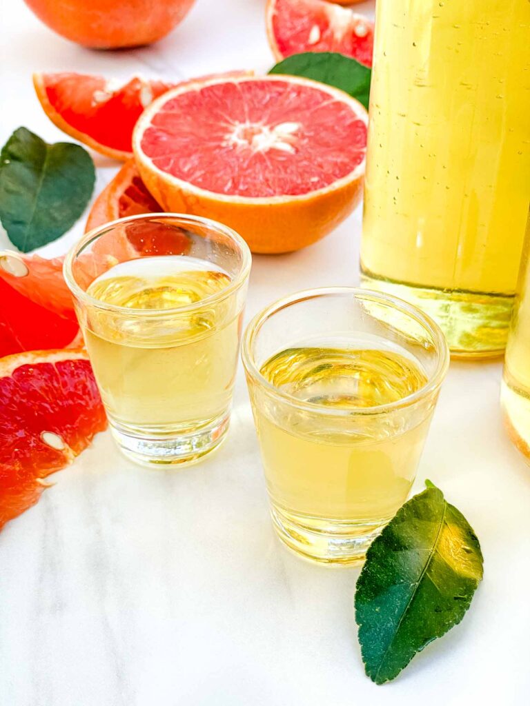 Two shot glasses of Grapefruit Limoncello (Grapefruit-cello) on a counter with fresh grapefruit around them.