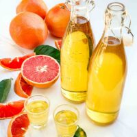 Two bottles of Grapefruit Limoncello (Grapefruit-cello) with two shots of it on a counter surrounded by fresh grapefruit.