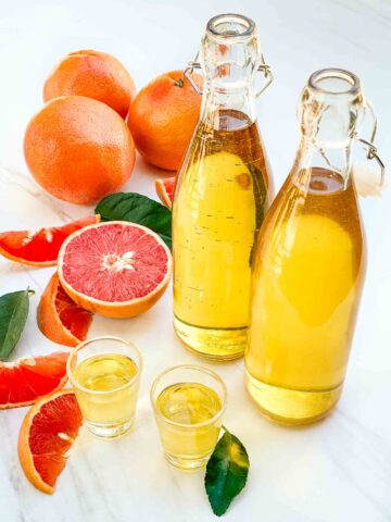 Two bottles of Grapefruit Limoncello (Grapefruit-cello) with two shots of it on a counter surrounded by fresh grapefruit.