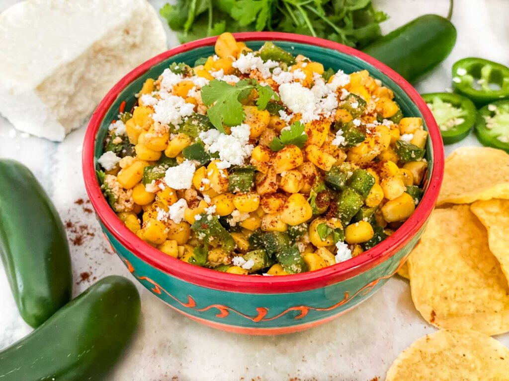 Mexican Street Corn Salsa (Esquites) in a colored bowl on a counter with peppers and chips around it