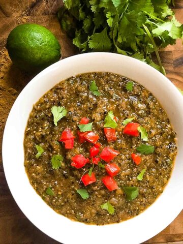 Roasted Tomatillo Salsa Verde in a white bowl with tomatoes and cilantro as a garnish. On a cutting board with cilantro and lime.