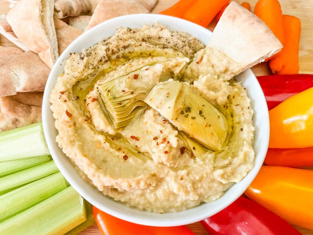 A bowl full of Artichoke Lemon Hummus surrounded by vegetables