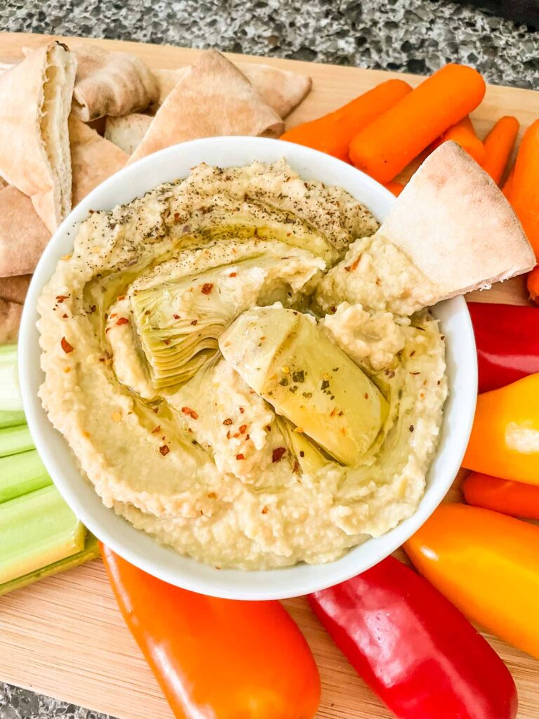 Top view of Artichoke Lemon Hummus in a small white bowl with vegetables around it