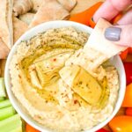 A hand dipping a piece of pita into a bowl of Artichoke Lemon Hummus