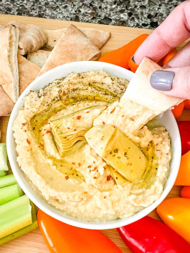 A hand dipping a piece of pita into a bowl of Artichoke Lemon Hummus