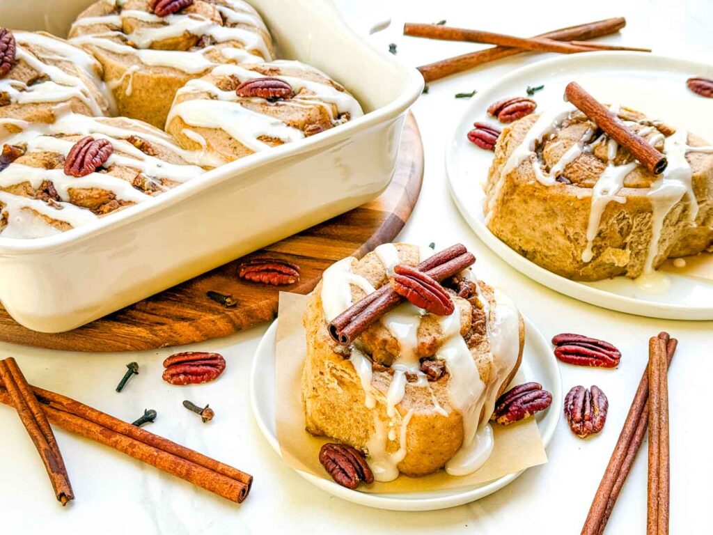 Pumpkin Spice Cinnamon Rolls with Maple Cream Cheese Glaze on small plates and in a baking dish, surrounded by nuts and cinnamon sticks.