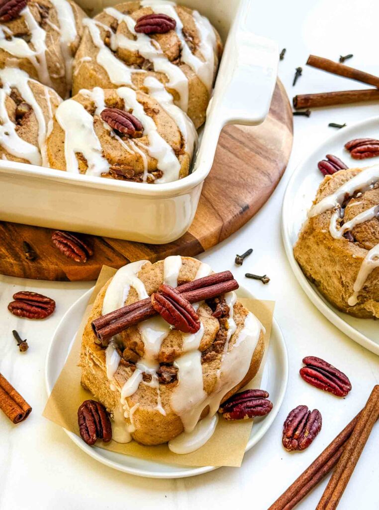 Some Pumpkin Spice Cinnamon Rolls with Maple Cream Cheese Glaze in a dish and on small plates, surrounded by pecans and cinnamon sticks.