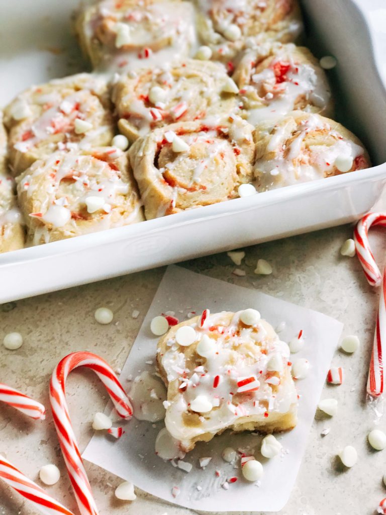 A white tray filled with Peppermint White Chocolate Sweet Rolls with one cinnamon roll on the counter