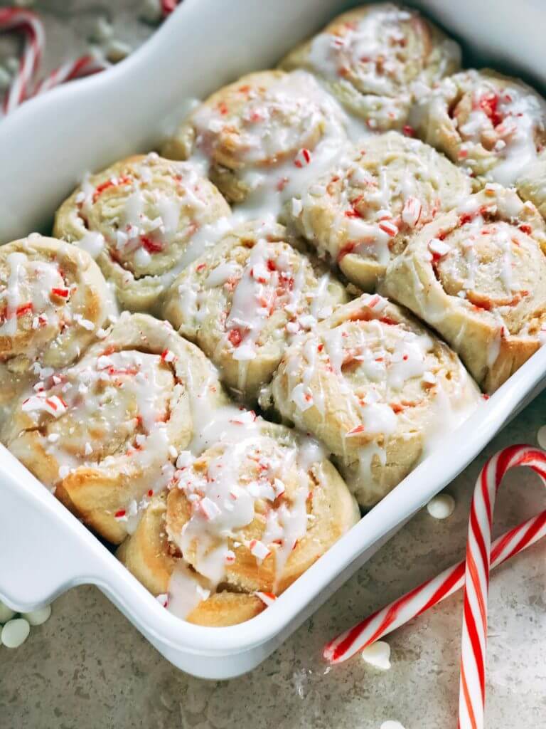 A white tray full of Peppermint White Chocolate Sweet Rolls with a white glaze surrounded by candy canes