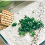 Pin of Ranch Hummus in a glass dish with fresh parsley on top and crackers. Title at top