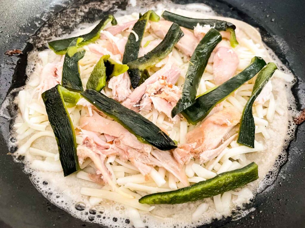 The tortilla with the cheese, chicken, and pepper on it cooking in a skillet before being folded