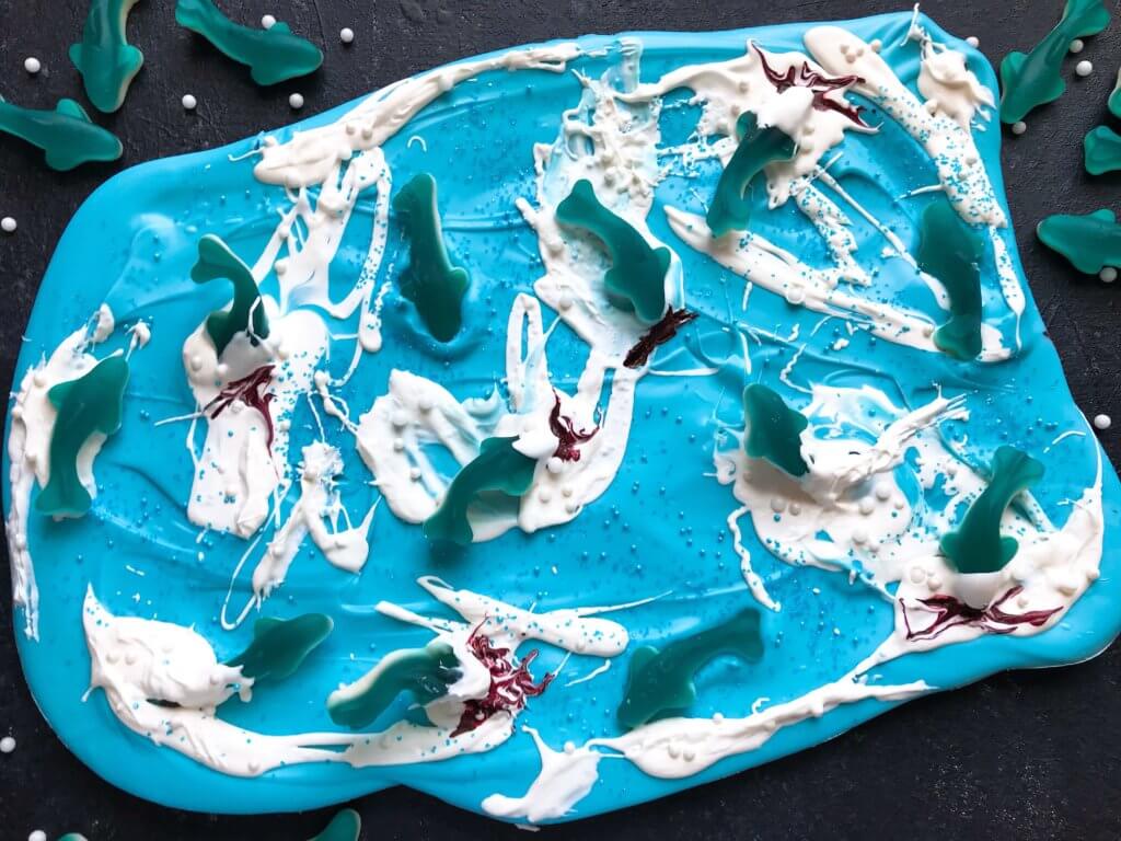 Just 10 minutes to make Shark Bark! Candy melts with sprinkles, gummy sharks, and a shark attack scene using red food gel. Gluten free (GF) quick and easy dessert recipe for shark or ocean party. Shark Week Shark Bark | Three Olives Branch | www.threeolivesbranch.com #sharkweek #ocean #sharkattack