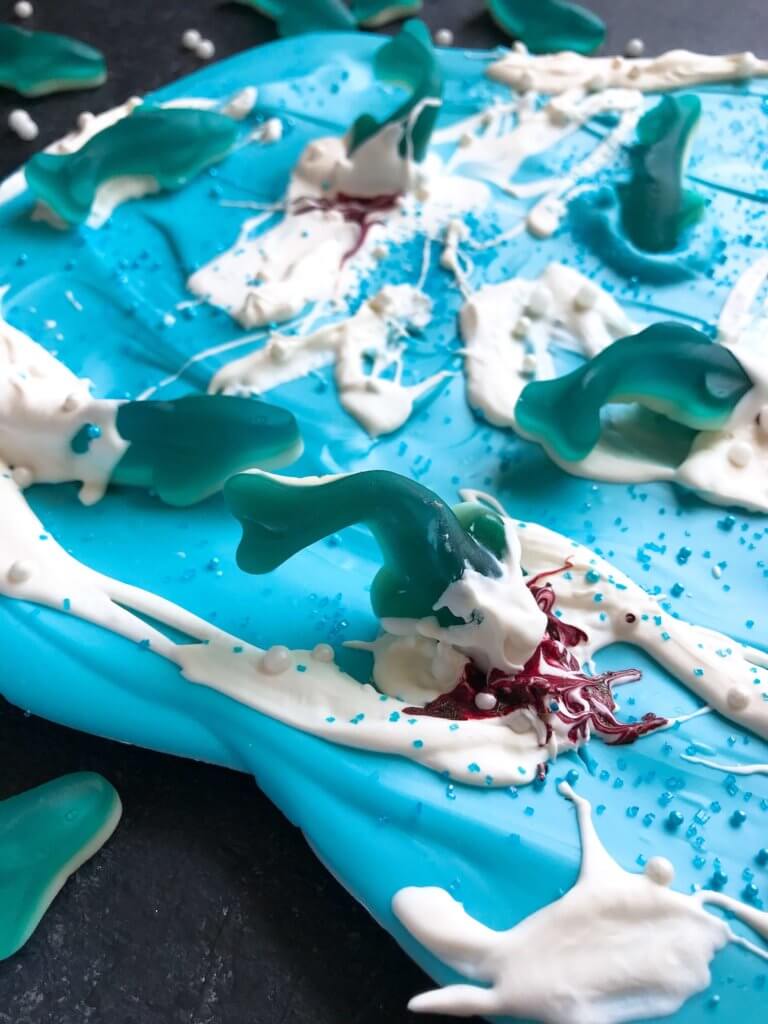Just 10 minutes to make Shark Bark! Candy melts with sprinkles, gummy sharks, and a shark attack scene using red food gel. Gluten free (GF) quick and easy dessert recipe for shark or ocean party. Shark Week Shark Bark | Three Olives Branch | www.threeolivesbranch.com #sharkweek #ocean #sharkattack