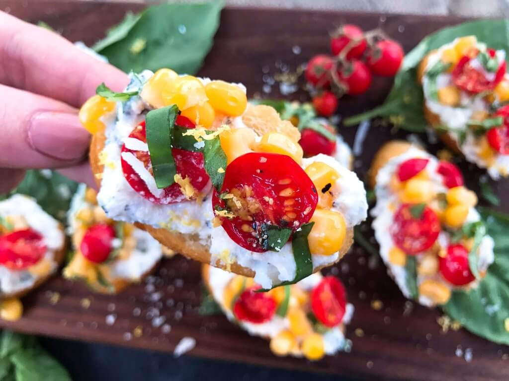 Fresh cherry tomatoes, basil, and corn are mixed with lemon. Ricotta and Parmesan cheese are mixed with lemon and herbs then layered on toasted bread slices. Tomato Corn Basil Crostini with Herbed Ricotta Cheese are vegetarian and perfect party and summer entertaining food. #fingerfood #summerappetizer #crostini