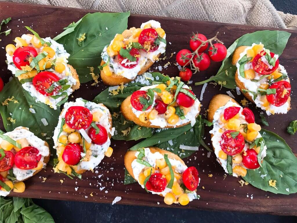 Fresh cherry tomatoes, basil, and corn are mixed with lemon. Ricotta and Parmesan cheese are mixed with lemon and herbs then layered on toasted bread slices. Tomato Corn Basil Crostini with Herbed Ricotta Cheese are vegetarian and perfect party and summer entertaining food. #fingerfood #summerappetizer #crostini