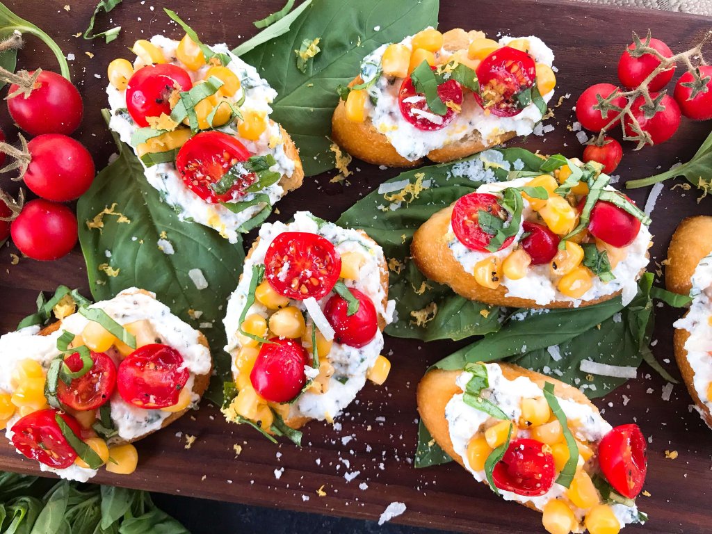 Fresh cherry tomatoes, basil, and corn are mixed with lemon. Ricotta and Parmesan cheese are mixed with lemon and herbs then layered on toasted bread slices. Tomato Corn Basil Crostini with Herbed Ricotta Cheese are vegetarian and perfect party and summer entertaining food. #fingerfood #summerappetizer #crostini