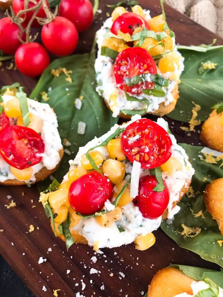 Fresh cherry tomatoes, basil, and corn are mixed with lemon. Ricotta and Parmesan cheese are mixed with lemon and herbs then layered on toasted bread slices. Tomato Corn Basil Crostini with Herbed Ricotta Cheese are vegetarian and perfect party and summer entertaining food. #fingerfood #summerappetizer #crostini