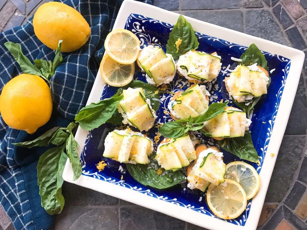A blue plate with Zucchini Lemon Ricotta Crostini surrounded by lemons and basil