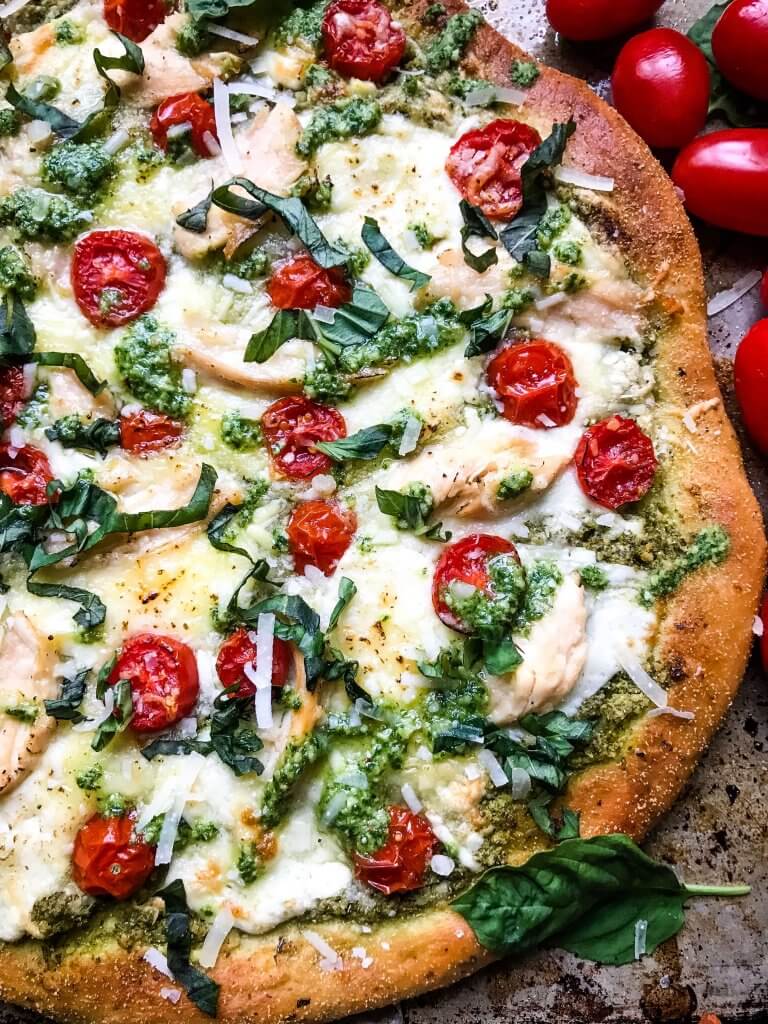 A whole Chicken Pesto Caprese Pizza on a baking sheet with extra tomatoes around it