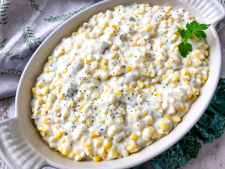 A white oval dish filled with creamy corn garnished with a sprig of parsley. The background features a light-colored cloth with green fern patterns.