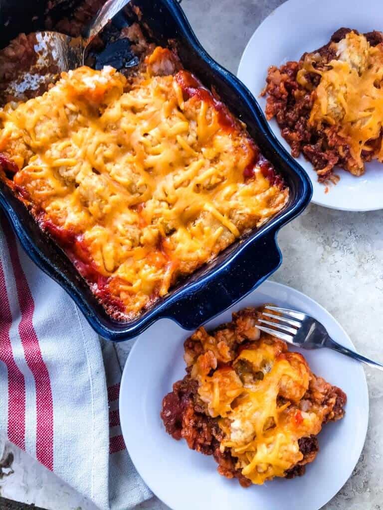 A dish full of Beef Sloppy Joe Tater Tot Casserole with two plates next to it that have a portion of the casserole on them