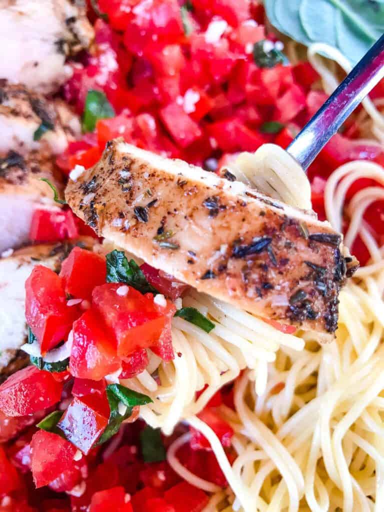 A fork scooping pasta with diced tomatoes and a slice of balsamic chicken