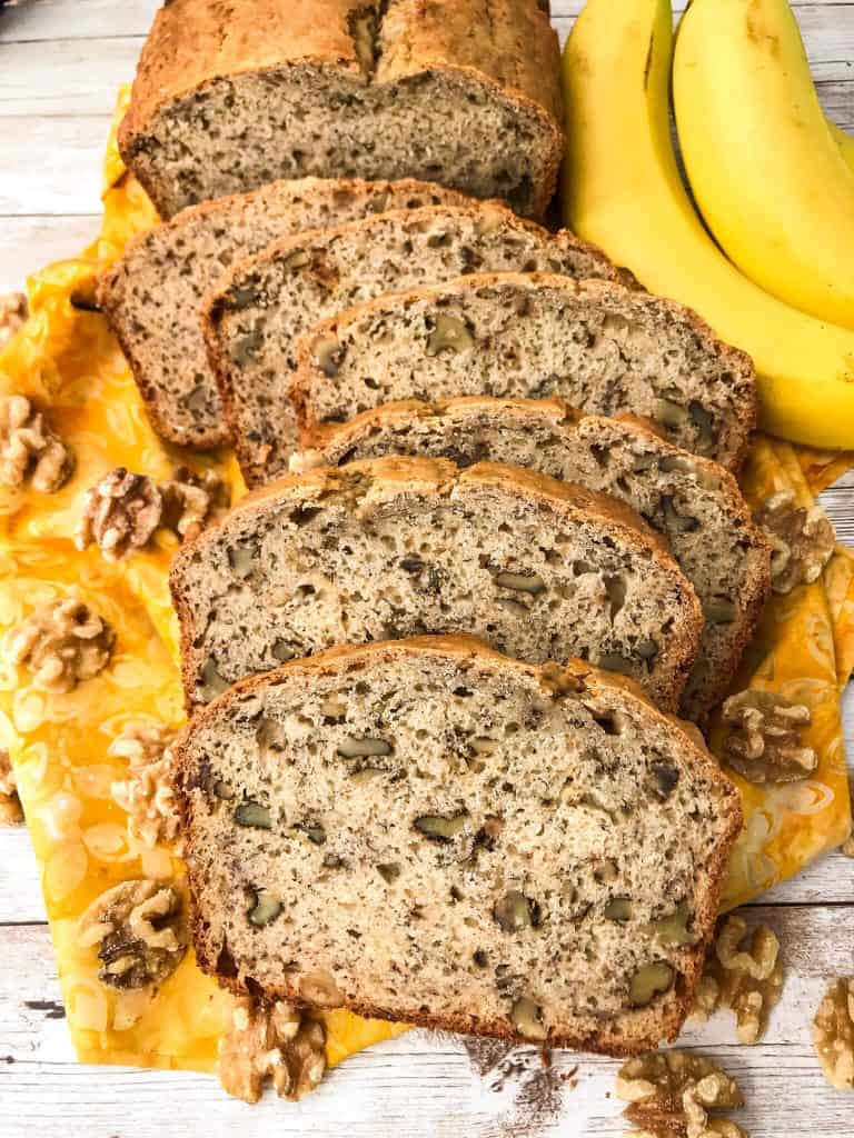 A loaf of Easy Banana Nut Bread cut in slices with bananas and walnuts next to it, on top of a yellow cloth