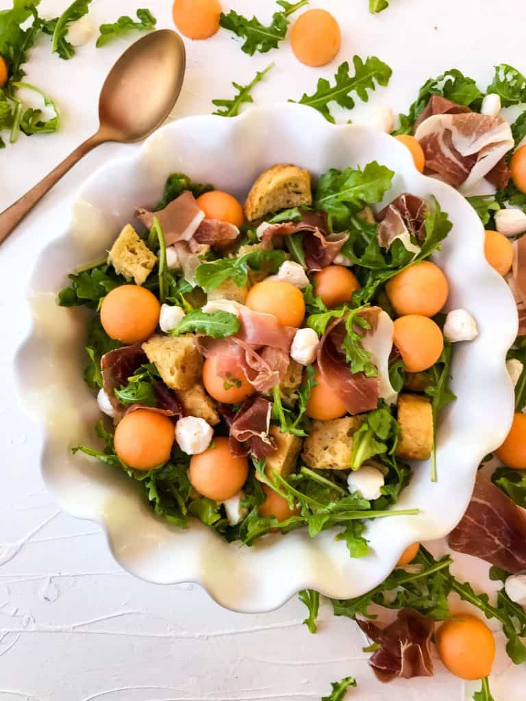 White bowl filled with Prosciutto Melon Panzanella Salad with a copper spoon and ingredients around it