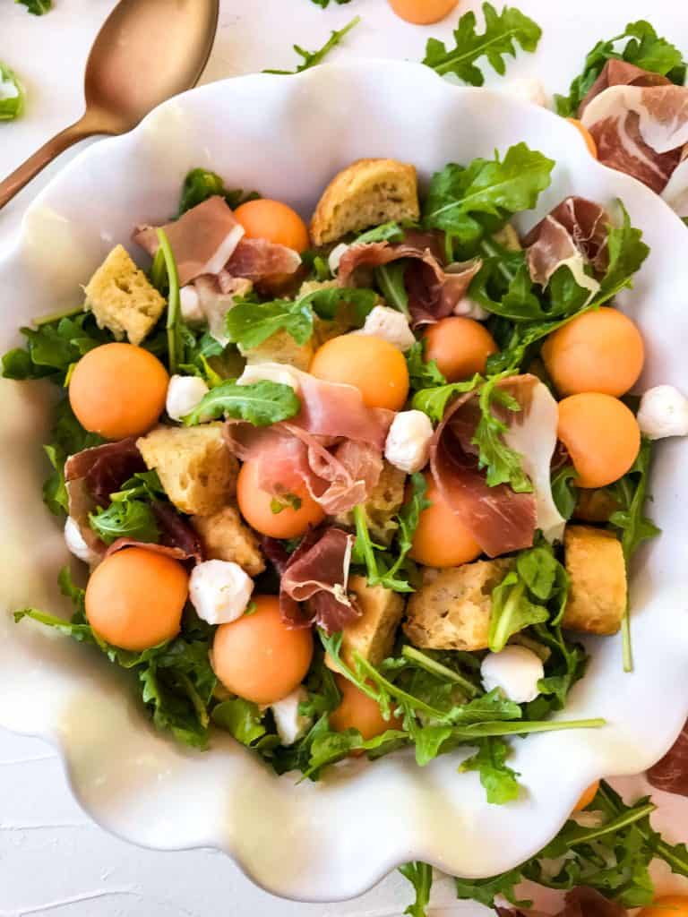 Cantaloupe, prosciutto, arugula, mozzarella, and bread salad in a white bowl