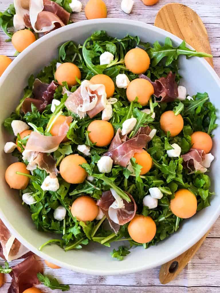 A bowl full of Prosciutto Melon Salad with Mozzarella and Arugula with a wood spoon