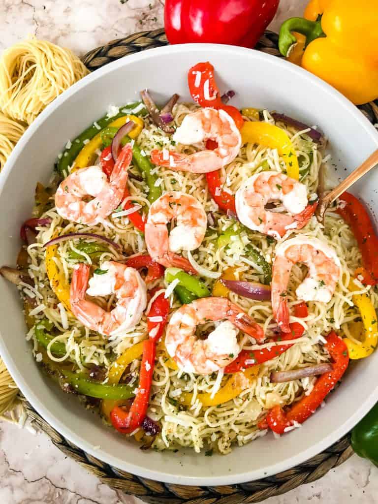 A bowl of Copycat Olive Garden Shrimp Scampi with a copper spoon and surrounded by bell peppers and fresh pasta