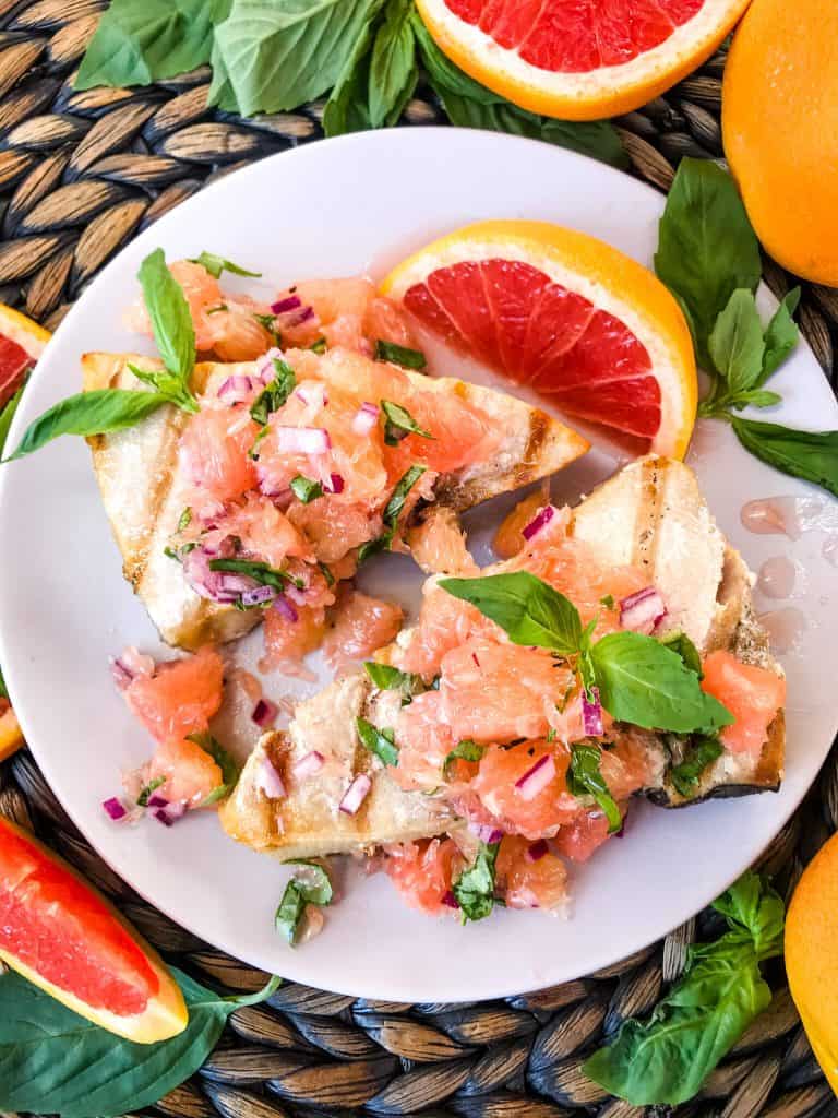 Overhead shot of two Basil Grapefruit Grilled Swordfish Steaks on a plate surrounded by grapefruit and basil