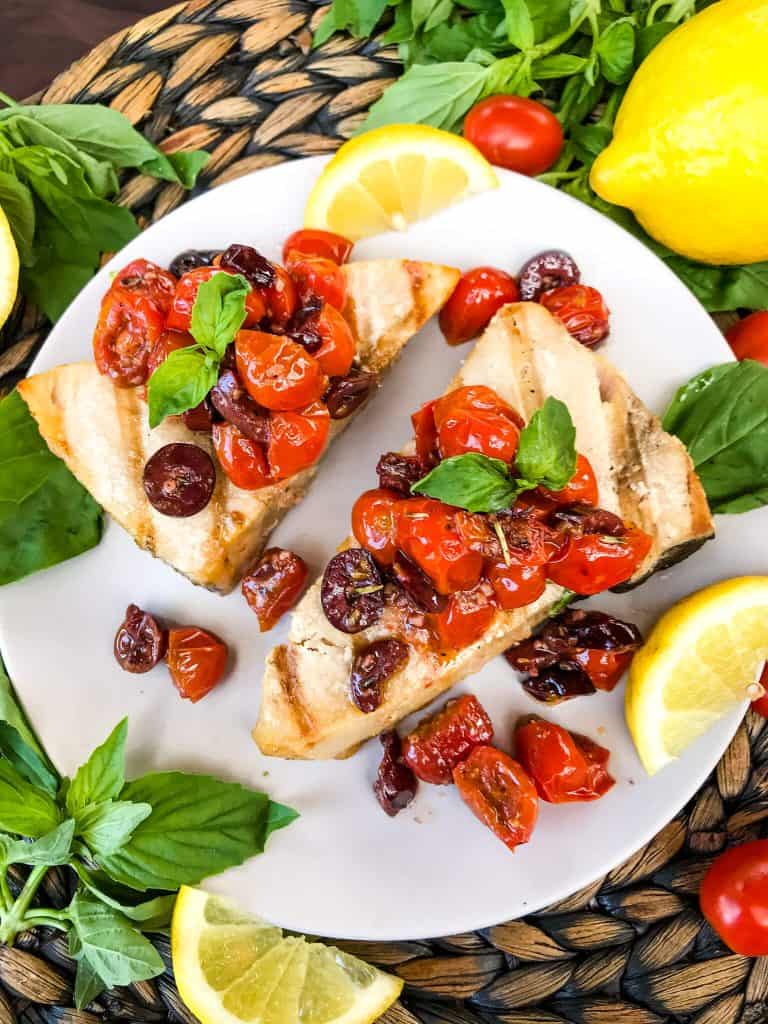 Two pieces of fish with tomato olive relish on top on a plate surrounded by lemons and basil