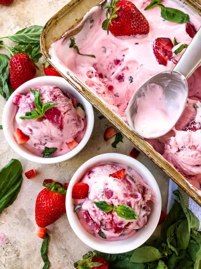 A pan full of No Churn Strawberry Basil Ice Cream with a scoop, and two small white bowls full of the ice cream next to it