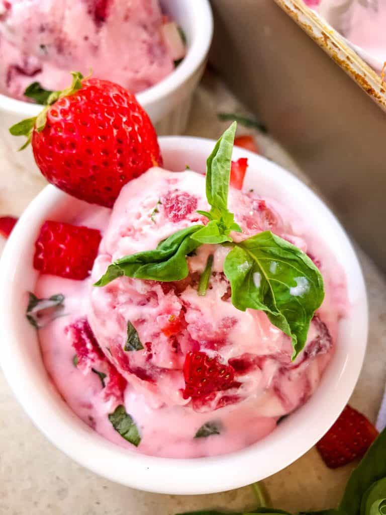 A scoop of strawberry basil ice cream in a small white bowl with a strawberry and fresh basil on top