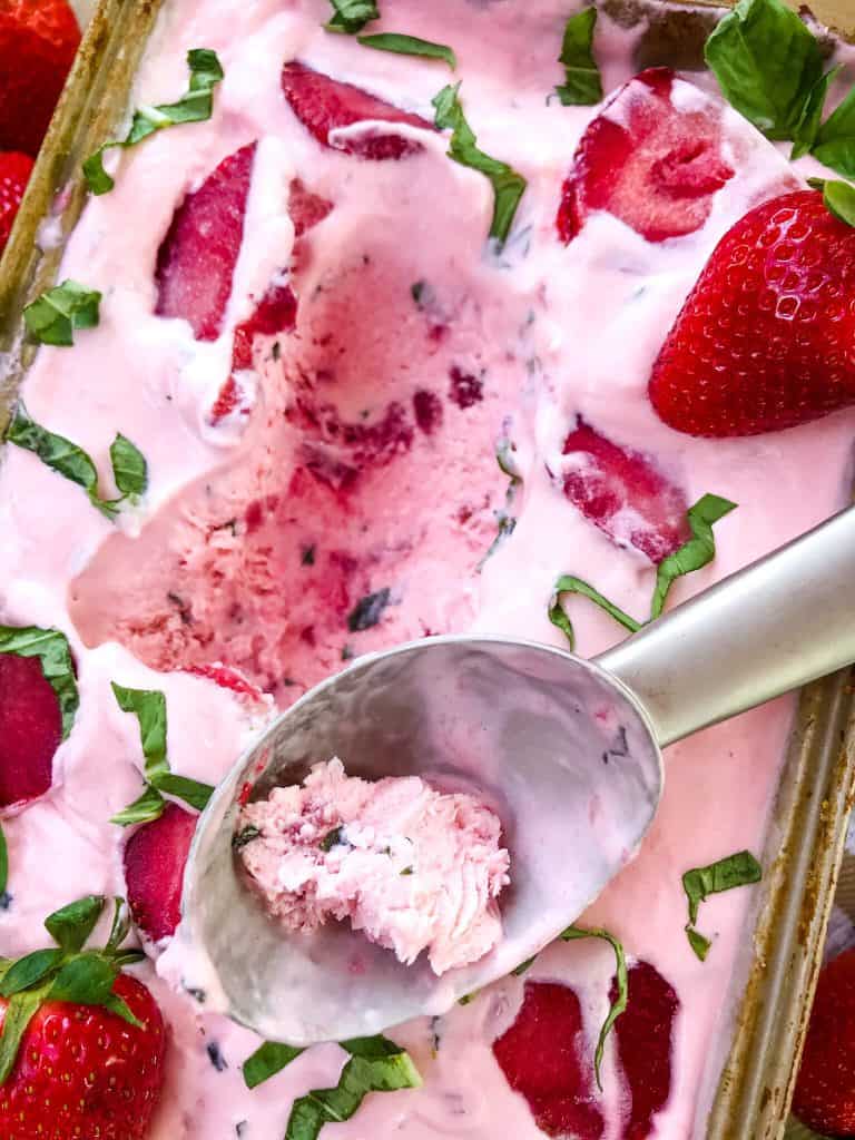 A pan full of No Churn Strawberry Basil Ice Cream with an ice cream scoop on top and a scoop of ice cream missing from the batch