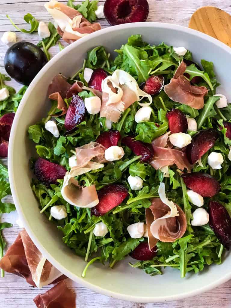 A bowl of Prosciutto Plum Arugula Salad with Mozzarella surrounded by ingredients and a wooden spoon