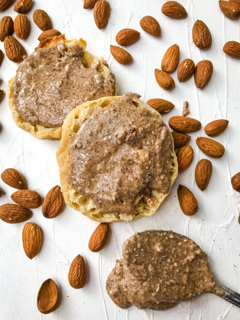 Two English muffins and a spoon with Homemade Crunchy Almond Butter Recipe surrounded by almonds