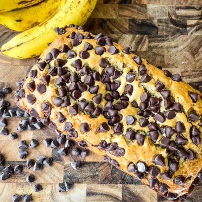 Top view of Chocolate Chip Banana Bread loaf on a wood cutting board next to chocolate chops and bananas