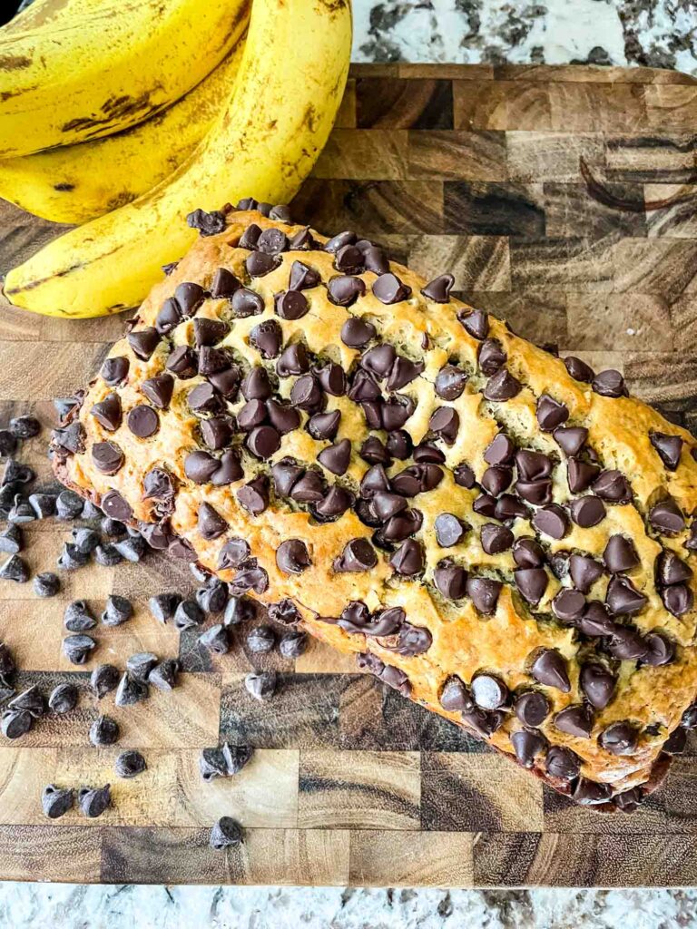 Top view of Chocolate Chip Banana Bread loaf on a wood cutting board next to chocolate chops and bananas