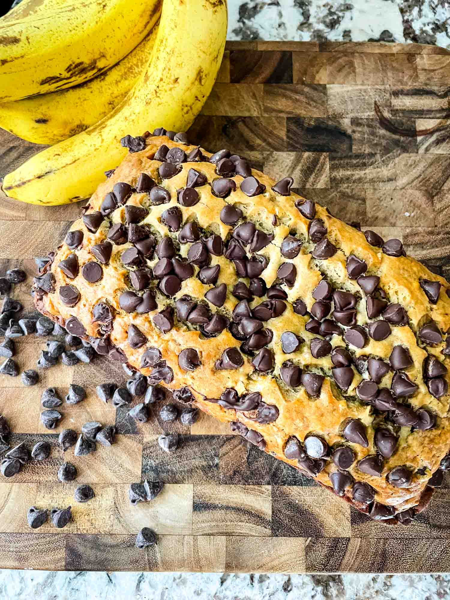 Top view of Chocolate Chip Banana Bread loaf on a wood cutting board next to chocolate chops and bananas