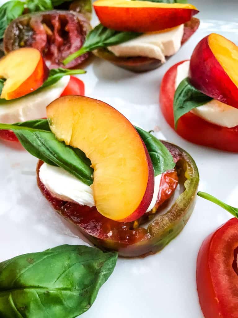 A stack of a tomato slice, fresh mozzarella, basil leave, and nectarine slice on white plate