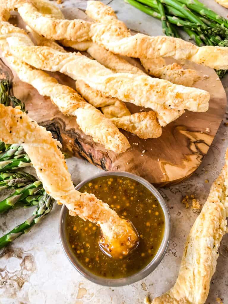 A cheese straw being dunked into honey mustard with a pile of Garlic Parmesan Cheese Straws